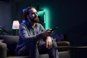 Man using remote control to switch channels, gripped by suspenseful films shown on television screen. Person mindlessly scrolling through streaming content, stopping after seeing interesting movie photo