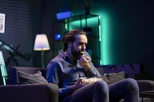 Lazy person lounged on couch using TV to binge series on subscription based streaming services, stuffing mouth with popcorn. Hungry man watching VOD shows on television display, eating unhealthy snack photo