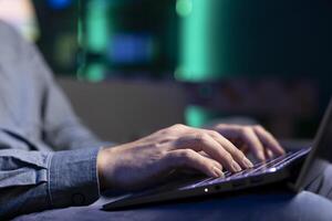 Close up shot of laptop used by man working from home in apartment typing on keyboard. Focus on notebook used by teleworking employee sending emails on couch in blurry background photo
