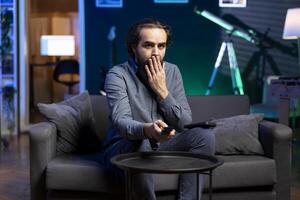 Man gasping while watching horror movie on streaming service, enjoying time off from work, holding remote control. Person covering mouth in disbelief, surprised by film scene broadcasted on TV photo