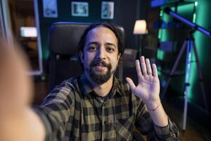 Male influencer waving his hand towards camera, making a daily vlog at home. Smiling young man having call with friends using modern technology. photo