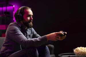 Man in neon lit apartment playing videogames on gaming console with teammates with bowl of popcorn in front. Player fighting foes in game, discussing with friends through headset and eating snacks photo