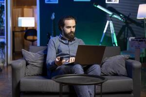 Man sitting in cozy apartment on couch typing on laptop keyboard and zapping through TV channels. Teleworking person solving tasks on notebook and picking film on streaming service photo