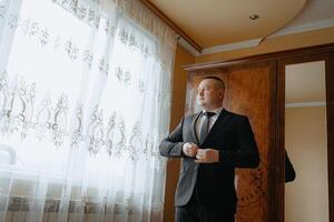 A man in a suit is standing in front of a window, adjusting his tie. Concept of formality and professionalism, as the man is dressed in a suit and tie photo
