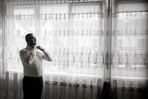 A man is getting dressed in front of a window. The curtains are white and the window is letting in some light. The man is wearing a white shirt and black pants photo