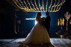 A bride and groom are standing in the rain, with the bride holding a bouquet. The scene is dark and moody, with the couple looking into each other's eyes photo