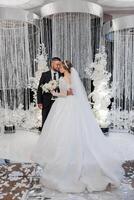 A bride and groom are standing in front of a white curtain with a waterfall. The bride is wearing a white dress and the groom is wearing a suit. They are holding hands and smiling at the camera photo