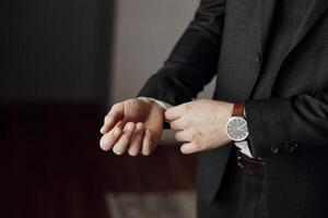A man in a suit is adjusting his watch. The man is wearing a black suit and a brown watch band photo