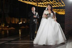 A bride and groom are posing for a picture in front of a building. The bride is wearing a white dress and holding a bouquet, while the groom is wearing a suit. The scene is set in a city photo