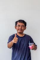 A young adult wearing a gray t-shirt and glasses is enjoying an Indonesian mixed iced drink or holding a plastic cup isolated on white background. for food content and health, and diabetes concepts photo