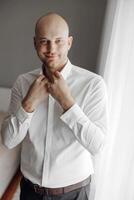 A man in a white shirt is standing in front of a window, adjusting his collar. The image has a casual and relaxed mood, as the man is getting ready for a day at home or work photo