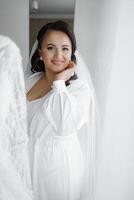 A woman in a white dress is posing for a picture. She is smiling and has her hand on her cheek. The image has a warm and friendly mood, and it seems like a happy moment captured in time photo
