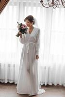 A bride is holding a bouquet of flowers and standing in front of a window. Scene is romantic and elegant, as the bride is dressed in a long white dress and is surrounded by the beauty of the flowers photo