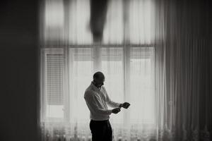 A man in a white shirt is standing in front of a window, adjusting his tie. The image has a moody and somewhat somber feel, as the man is in a reflective or contemplative state photo