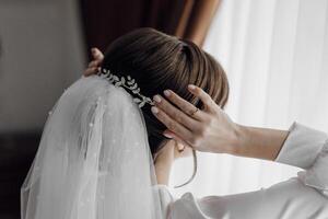 A woman is getting ready for a wedding and is putting on a veil. The veil is made of lace and has a leaf design. The woman is standing in front of a window, and the sunlight is shining on her face photo