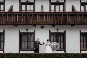 A bride and groom are standing on a balcony of a building. The bride is wearing a white dress and the groom is wearing a suit. They are holding hands and smiling at the camera photo