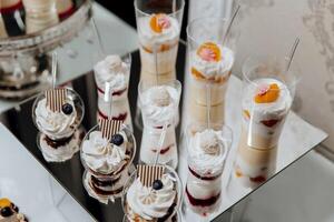 A table with a variety of desserts in clear plastic cups. The desserts include a blueberry cake, a strawberry shortcake, and a vanilla pudding. The table is set up for a party or gathering photo