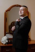A man in a suit is standing in front of a mirror, adjusting his tie. Concept of formality and attention to detail, as the man takes the time to ensure his appearance is polished and professional photo