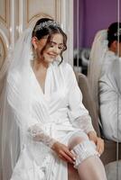 A bride is sitting in a white robe and wearing a veil. She is smiling and she is happy photo