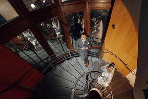 A bride and groom are walking down a spiral staircase. The bride is wearing a white dress and the groom is wearing a suit. The scene is set in a large room with many people in the background photo