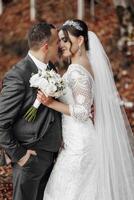 A bride and groom are hugging and kissing each other. The bride is wearing a white dress and a veil. They are standing in a field with leaves on the ground photo
