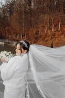 A woman in a white jacket is holding a bouquet of flowers. She is wearing a veil and a headband. The scene is set in a forest with trees in the background photo