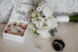 A bouquet of white flowers sits on a table next to a white box and a watch. The flowers are arranged in a vase and the box contains a pair of earrings photo