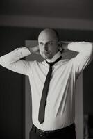 A man in a white shirt and black tie is standing in front of a door. He is adjusting his tie and looking at the camera. Concept of formality and professionalism, as the man is dressed in a suit photo