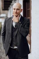 A man in a suit and vest is standing in front of a building. He is wearing a watch and a green flower in his hand. Concept of formality and elegance, as the man is dressed in a suit photo