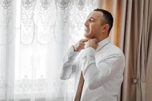 A man is getting ready for work, putting on a white shirt and a tie. He is smiling, indicating that he is happy and confident about his appearance photo