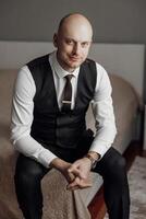 A man in a suit and vest sits on a bed. He is smiling and wearing a tie photo