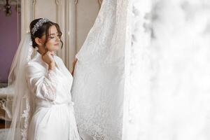A woman in a white gown is standing in front of a window, holding a white dress. The dress is draped over a curtain, and the woman is adjusting it. Concept of elegance and sophistication photo