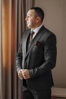 A man in a suit and tie stands in front of a window. He is wearing a red rose tie and a black suit photo
