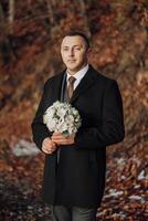 A man wearing a black coat and tie holding a bouquet of flowers. The man is standing in a forest with a backdrop of trees photo