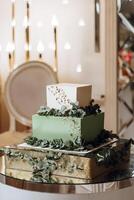 A three tier cake with green leaves on top. The cake is on a table with a chair in the background photo