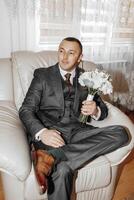 A man in a suit and tie is sitting in a chair holding a bouquet of white flowers photo