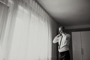 A man is getting ready for work, putting on his tie and adjusting his collar. The room is dimly lit, with a window letting in a small amount of light photo