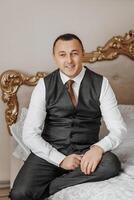 A man in a suit and vest sits on a bed. He is smiling and he is happy photo