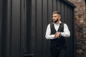 A man in a black vest and white shirt is standing in front of a brick wall. He is wearing a watch and he is checking the time photo