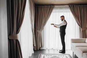 A man in a suit is standing in front of a window, adjusting his tie. The room is decorated with curtains and a rug, giving it a cozy and comfortable atmosphere photo