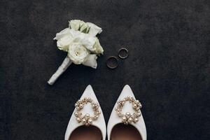 A pair of white shoes with a pearl embellishment and a boutonniere. The shoes are placed on a dark surface, and the boutonniere is next to them. Concept of elegance and sophistication photo