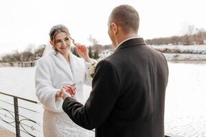 A bride and groom are standing on a bridge near a body of water. The bride is wearing a white coat and the groom is wearing a black suit. They are holding hands and smiling at the camera photo