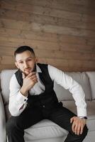 A man in a suit and vest sits on a couch, looking thoughtful. He is wearing a watch and has a beard photo