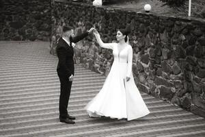 A bride and groom are dancing on a stone walkway. The bride is wearing a long white dress and the groom is wearing a suit. Scene is joyful and celebratory, as the couple is enjoying their wedding day photo