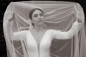 A woman is wearing a white dress and veil, and is standing in front of a white curtain photo