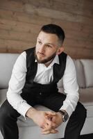 A man in a suit and vest sits on a couch. He is wearing a watch and has a beard photo