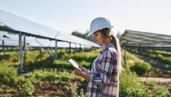 Beautiful female engineer wearing a white helmet and glasses monitoring solar panels on a tablet.Generated image photo