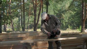 An elderly man works diligently, using a circular saw to cut wooden planks in a forest. Surrounded by tall trees, he demonstrates craftsmanship with precision and care, while focused on his task. video