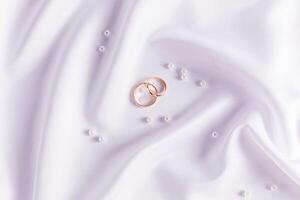 Two gold wedding rings on a white satin background with soft waves of textiles among the pearls. top view. wedding background. photo