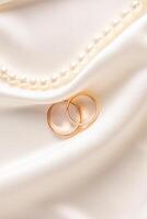 Two classic gold wedding rings lie among the delicate folds of satin fabric with a string of pearl beads. vertical front view. postcard, invitation. photo